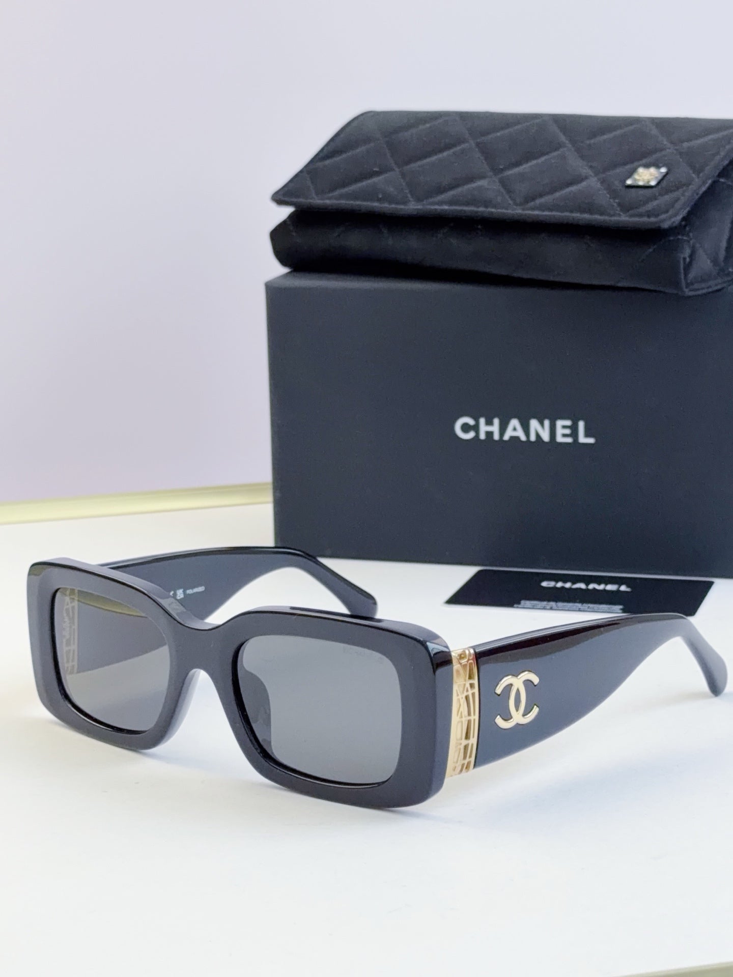 Square sunglasses with dark gray frames and tinted lenses, gold logo detail