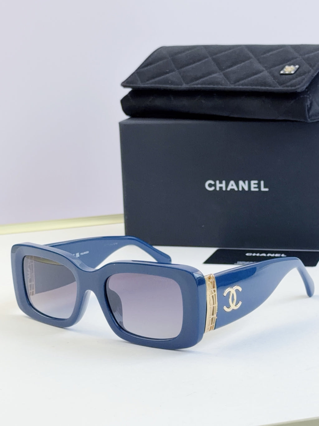 Square sunglasses with blue frames and tinted lenses, gold side logo detail