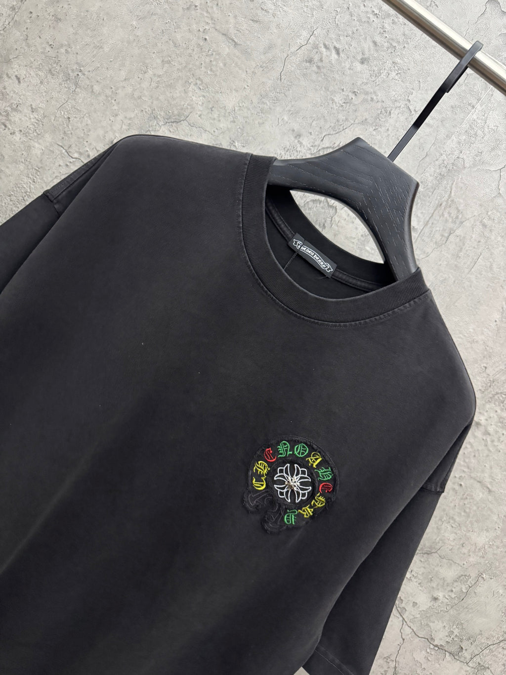 Black crewneck T-shirt close-up with small multicolor flower logo embroidery.