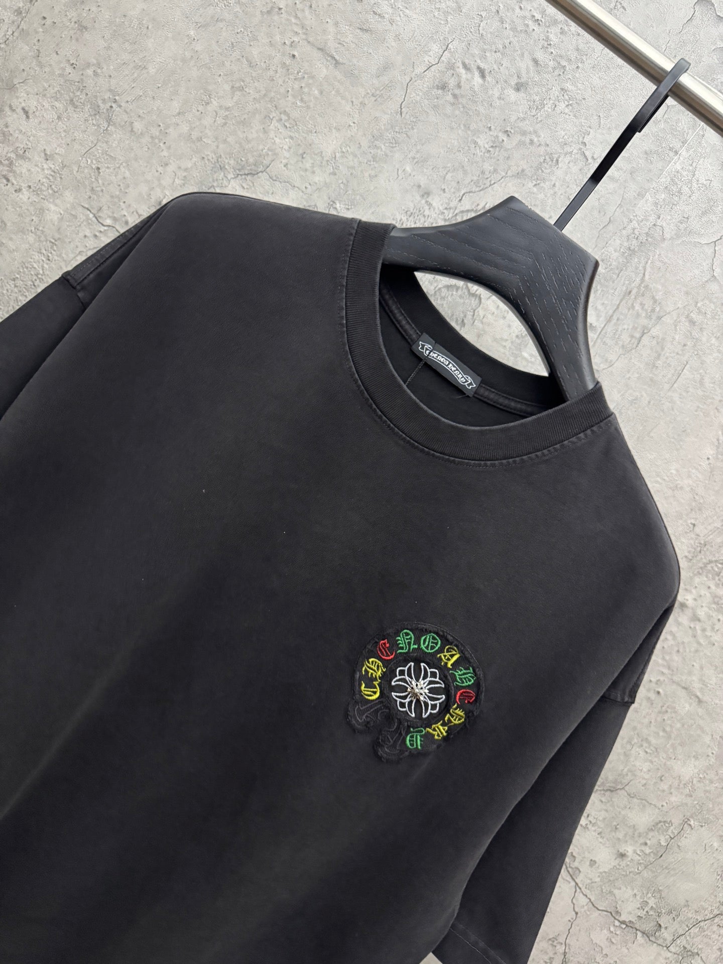 Black crewneck T-shirt close-up with small multicolor flower logo embroidery.