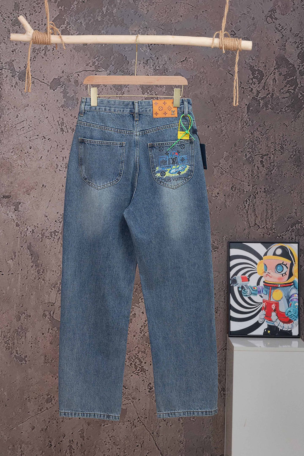 Blue denim pants with Louis Vuitton graphic patch on back pocket, hanging on wooden hanger.