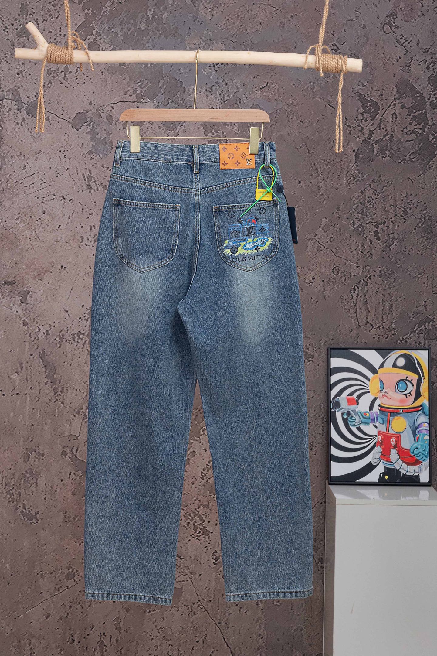 Blue denim pants with Louis Vuitton graphic patch on back pocket, hanging on wooden hanger.