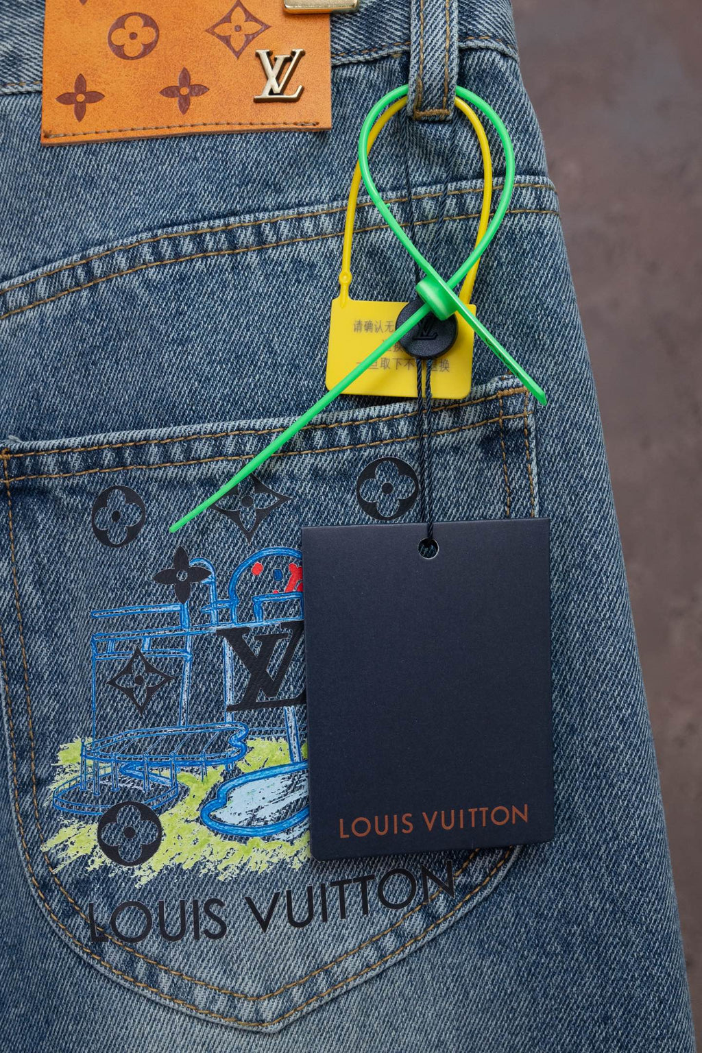 Blue denim jeans with printed graphic patch and attached Louis Vuitton hang tag.