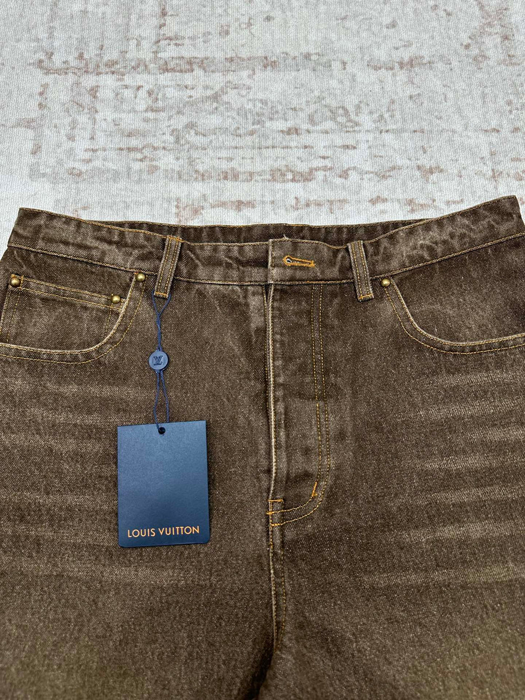 Brown denim jeans close-up with curved front pockets, gold buttons, and hanging tag