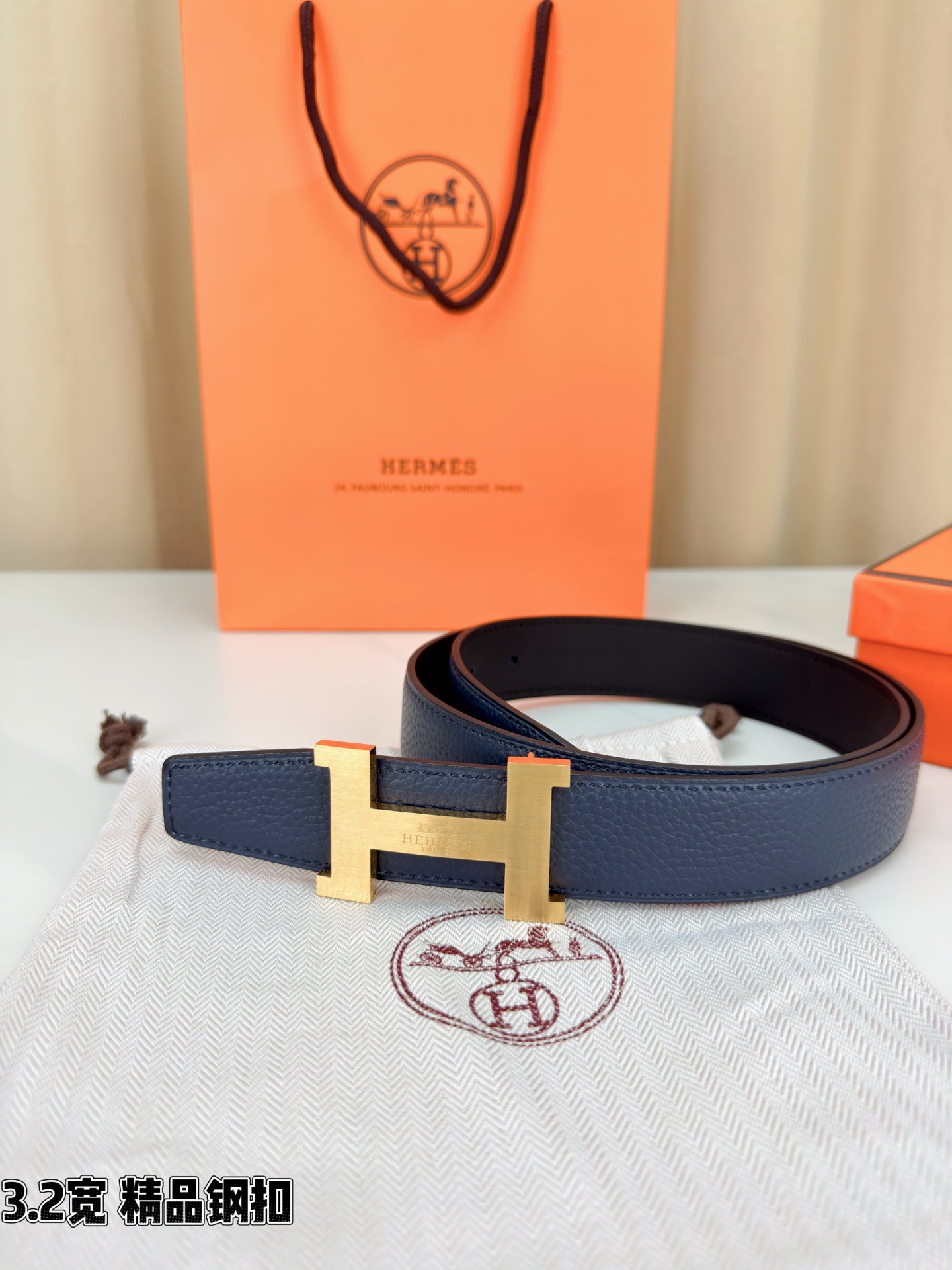 Dark blue leather belt with gold H buckle, draped over a light fabric pouch