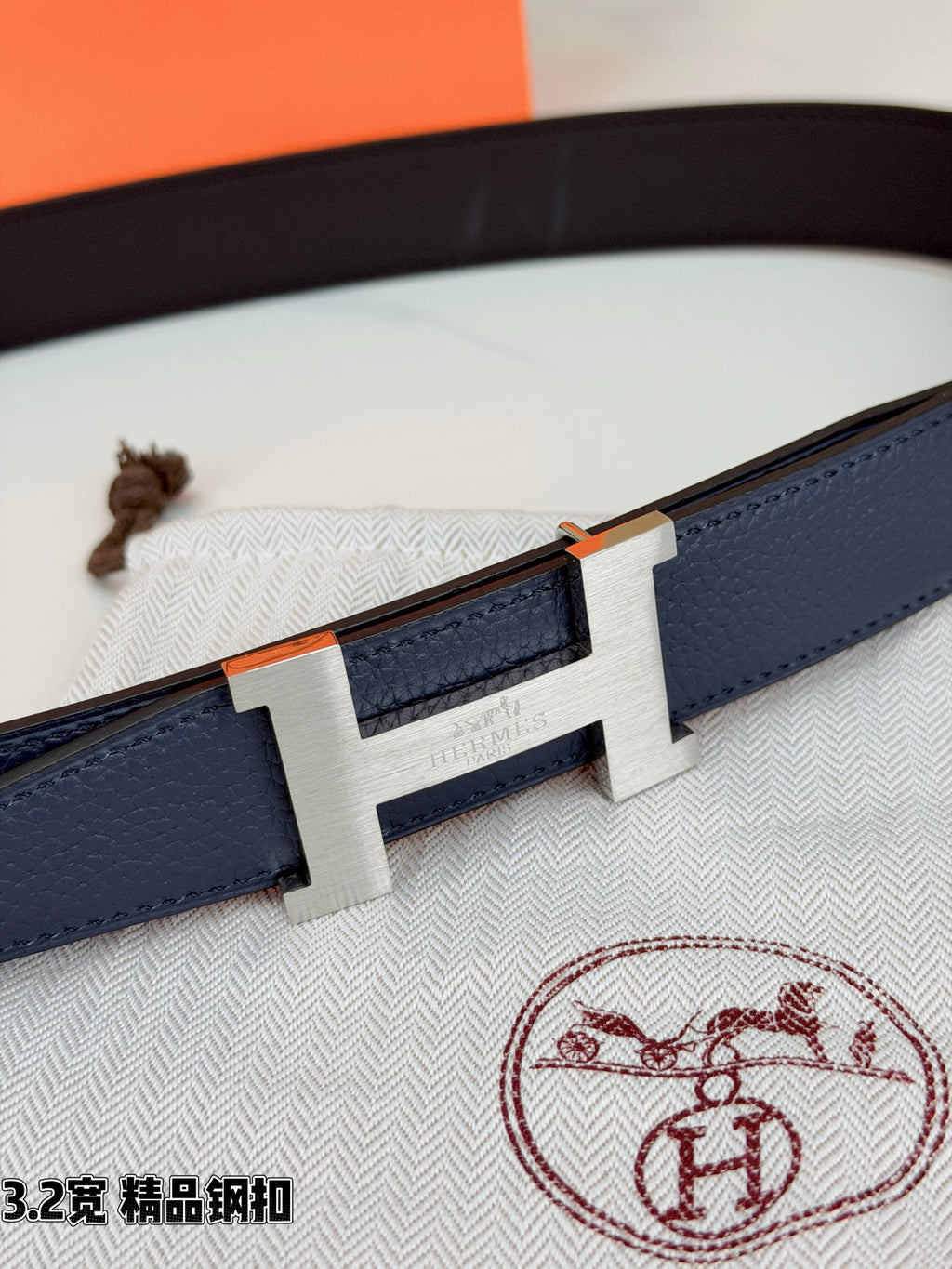 Close-up of dark blue leather belt with rectangular gold H buckle and engraved center