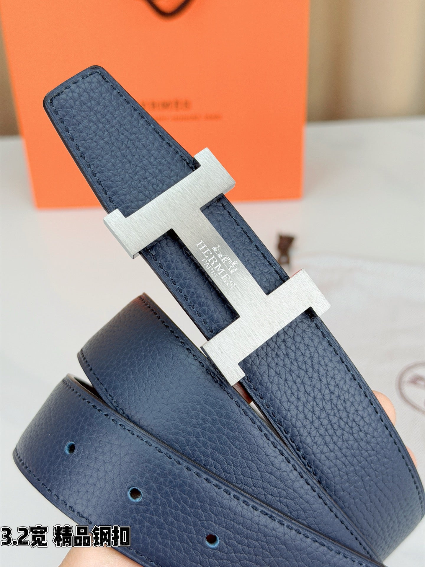 Dark blue leather belt with gold H buckle, angled view showing textured strap