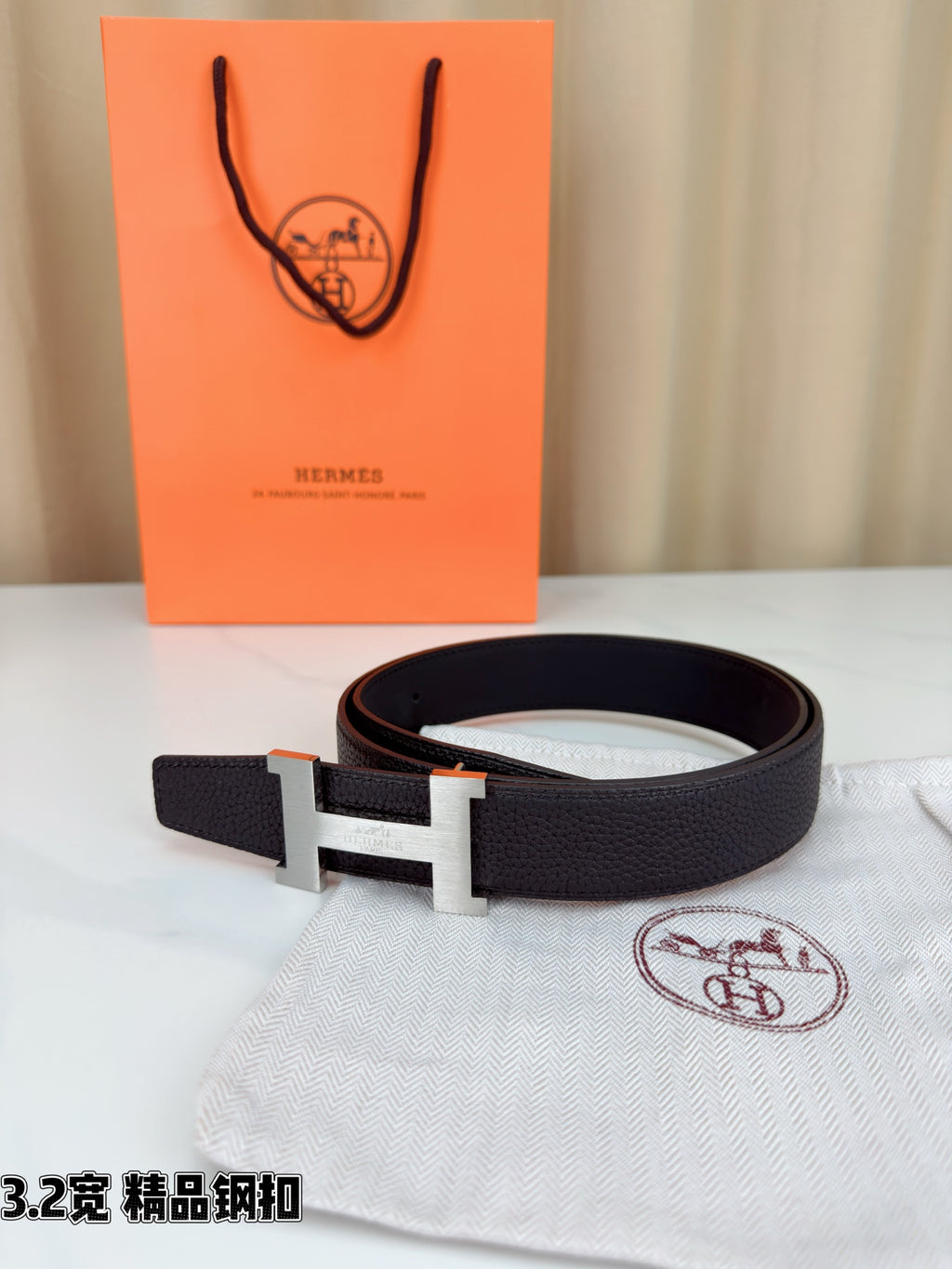 Leather belt with dark strap and gold-tone H buckle, laid across fabric surface.