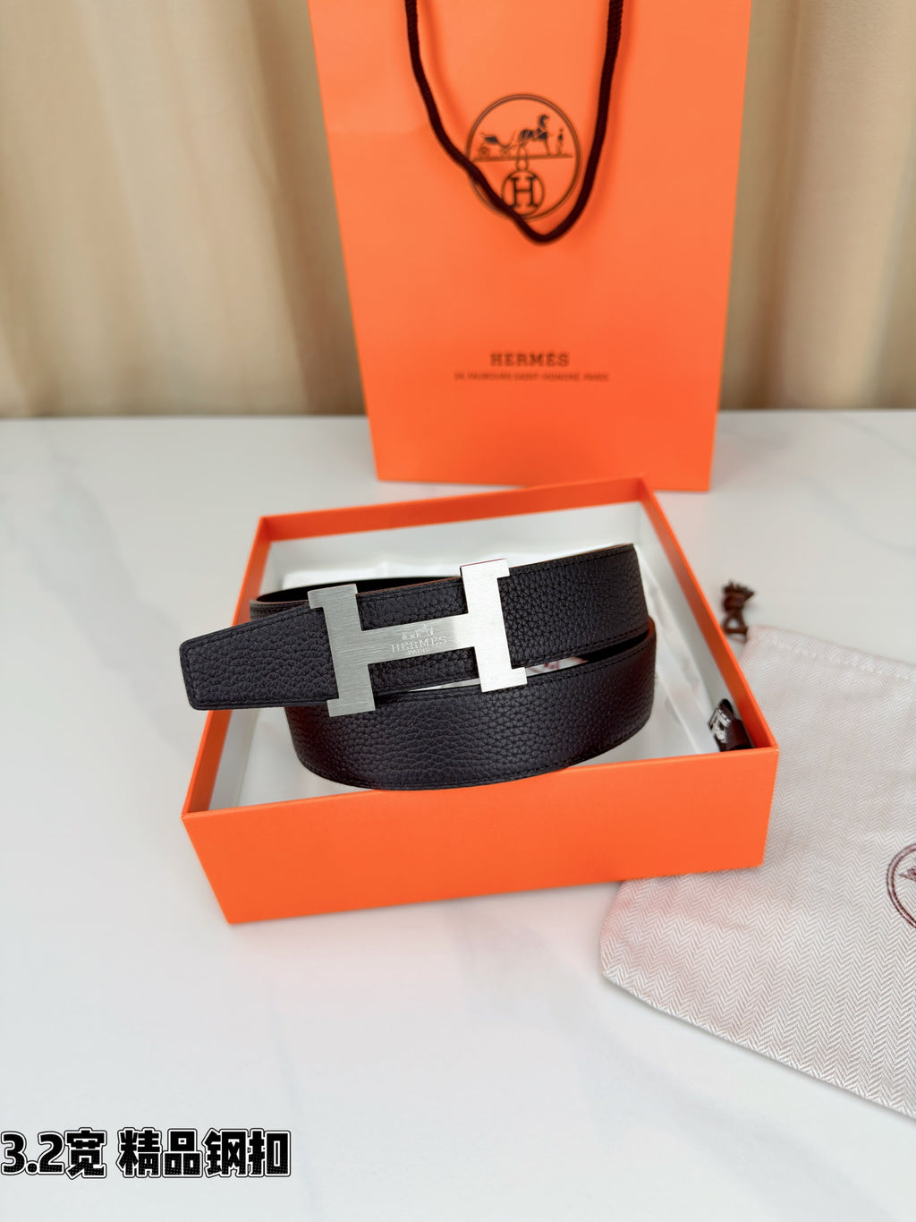 Leather belt with dark textured strap and gold-tone H buckle, resting in open box.