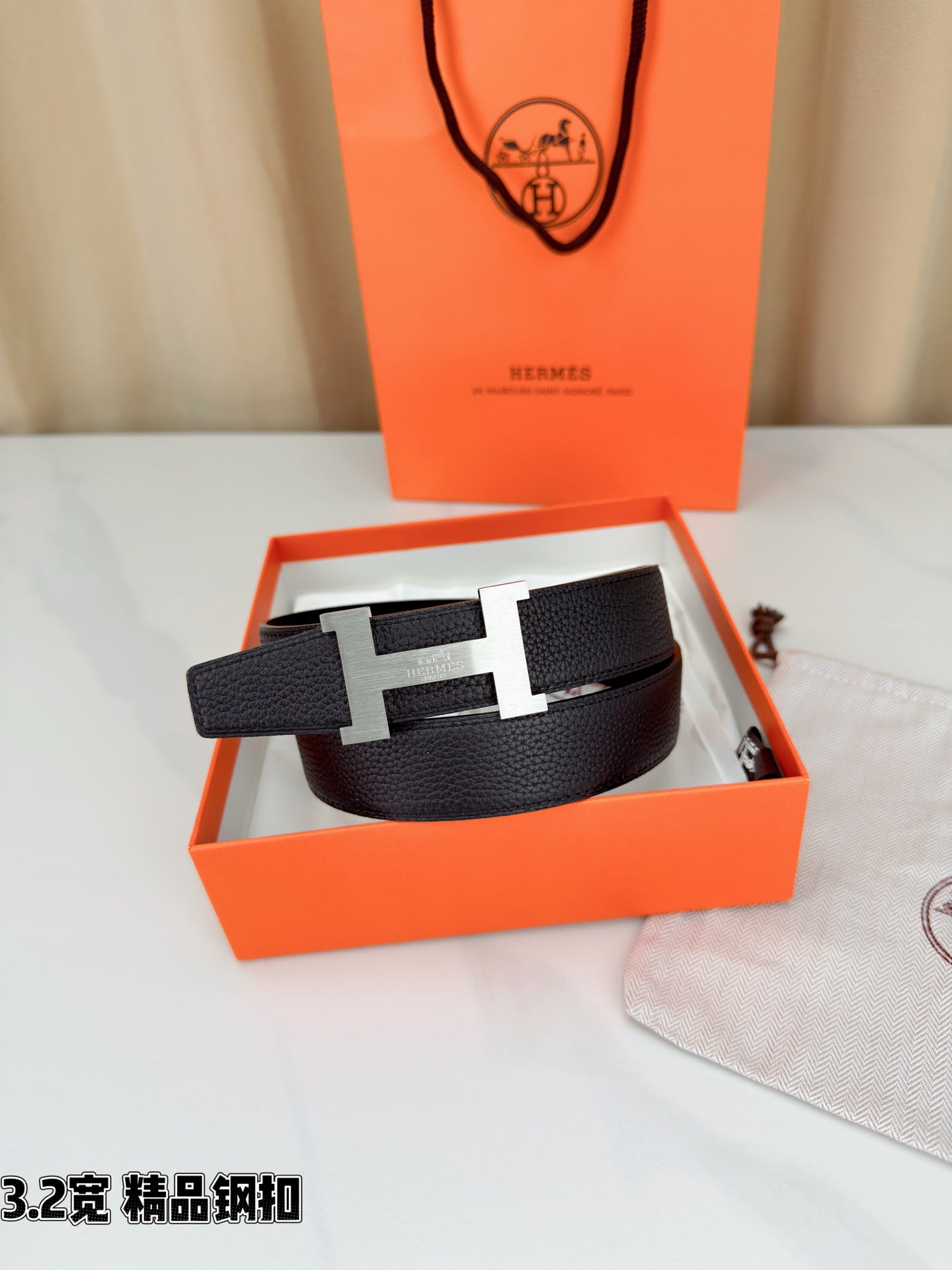 Leather belt with dark textured strap and gold-tone H buckle, resting in open box.