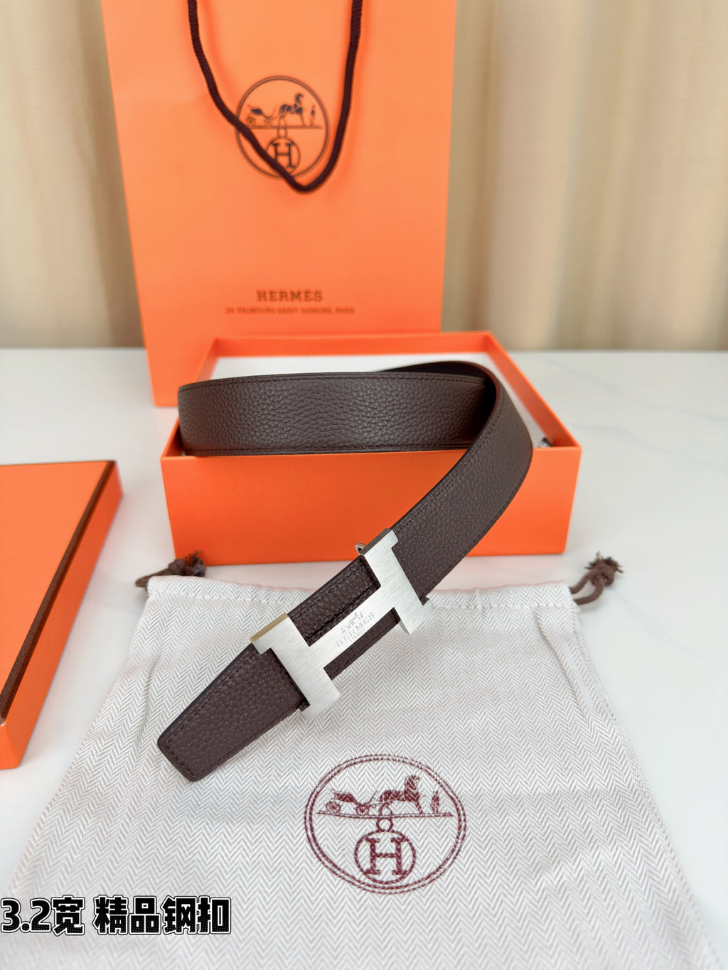 Dark brown leather belt with gold-tone H buckle on white dust bag.