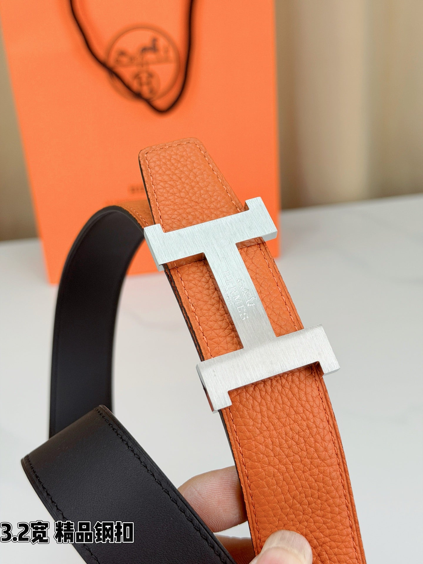Orange leather belt with gold-tone H buckle detail, held in hand