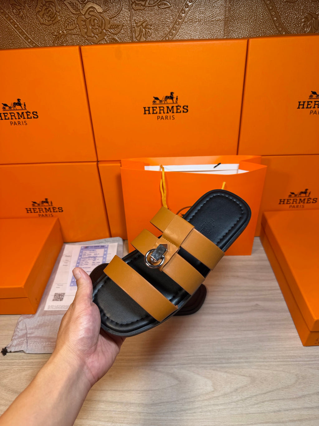 Hermes sandals held in hand: tan strap, dark footbed, silver buckle.