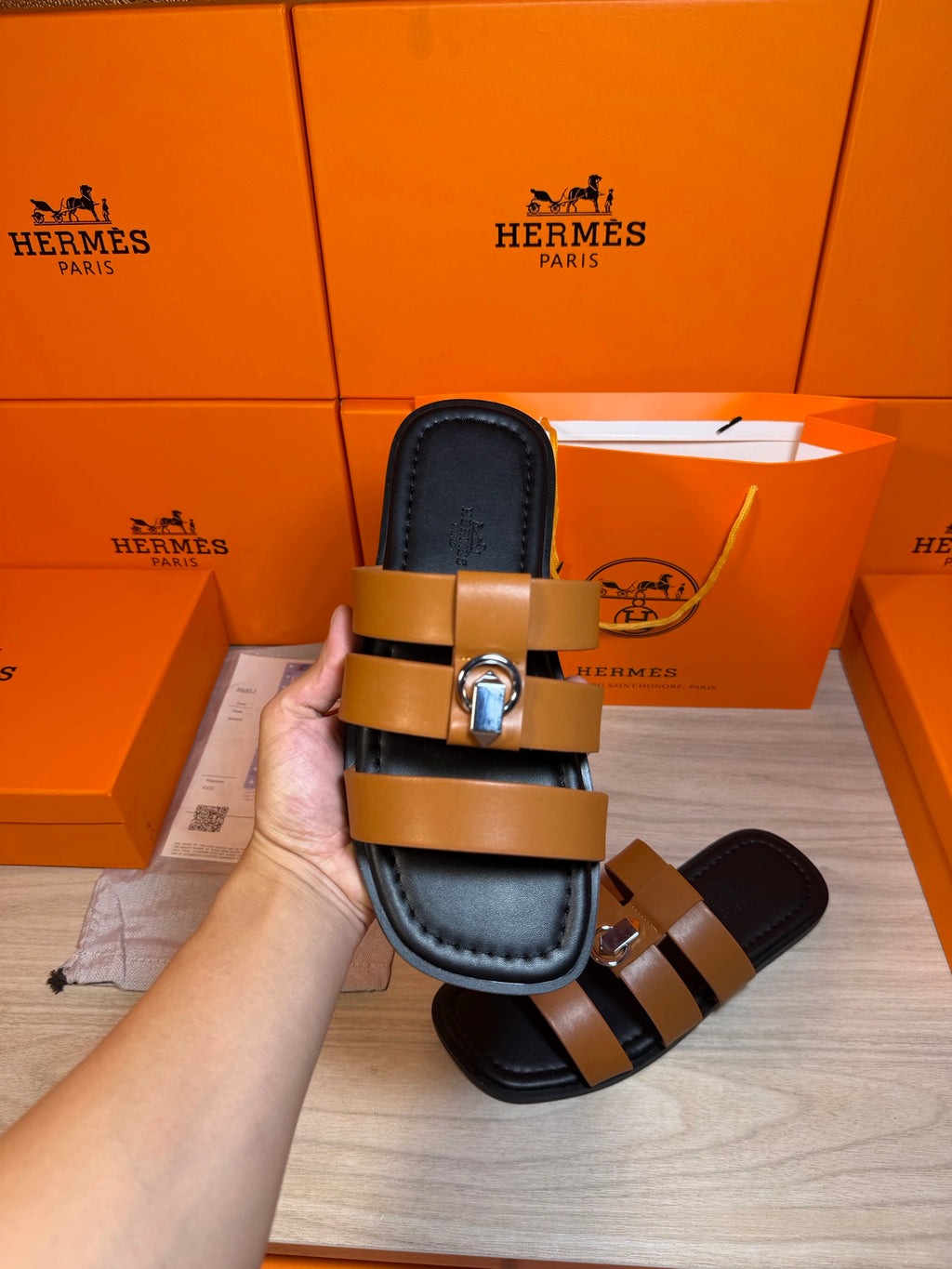 Hermes sandal close-up: tan strap bands over dark footbed with silver buckle.