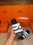 Luxury Hermes slide sandal held up: white straps over black base, silver buckle detail.