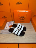 Luxury Hermes slide sandals with two white straps and silver buckle on black footbed.