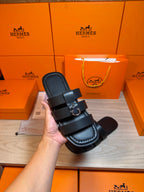 Hand holding dark leather Hermes slide sandal with two straps and metal buckle