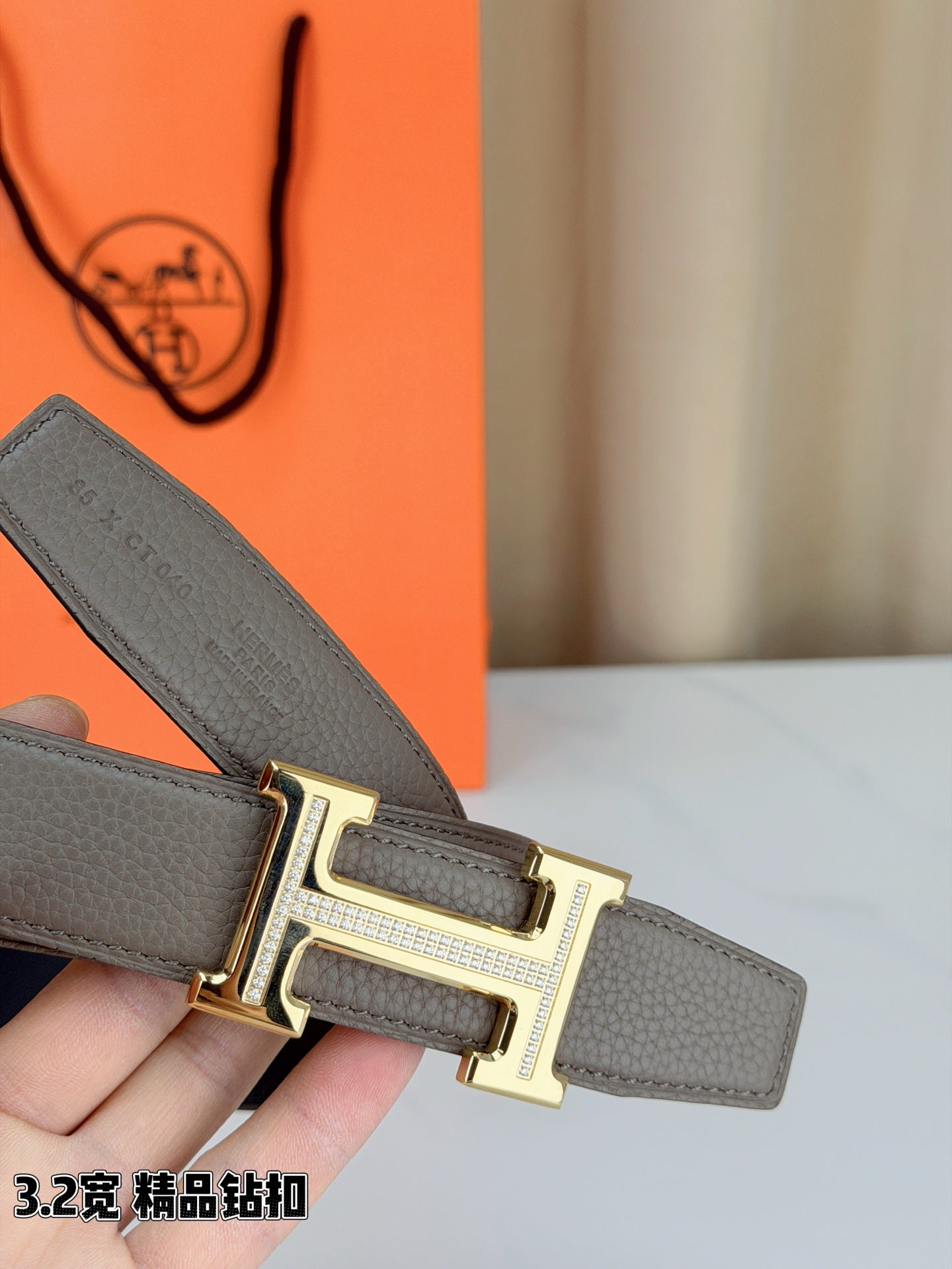 Light gray leather belt with gold-tone H buckle fastening, close-up view