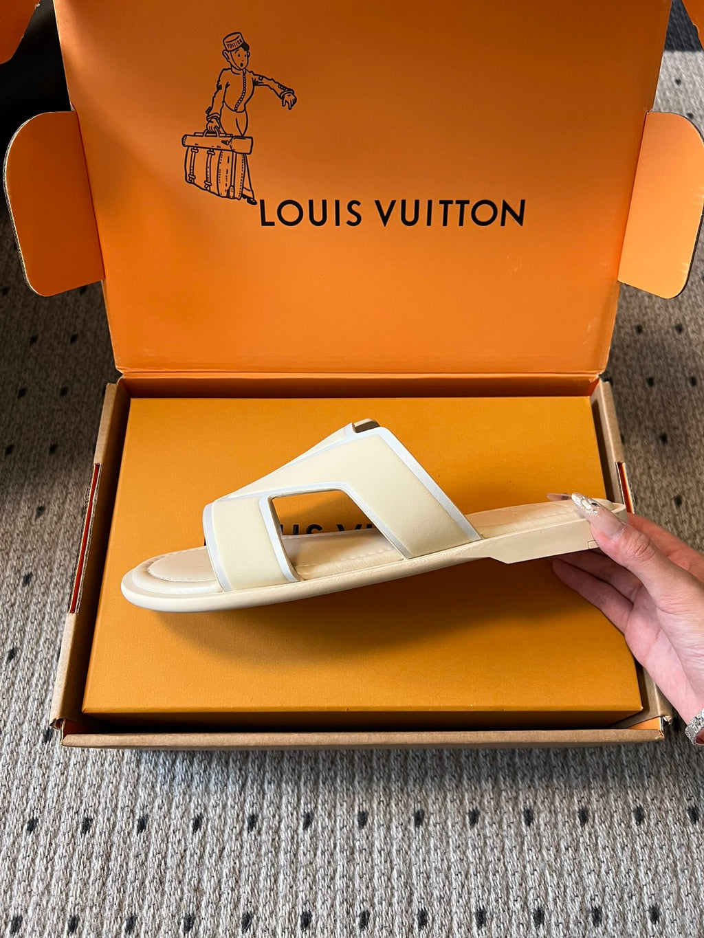 Cream slide sandal held above open box, V-shaped strap and cushioned footbed