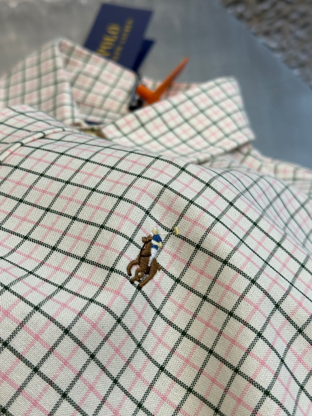 Plaid shirt fabric close-up with embroidered horse logo and product tags.