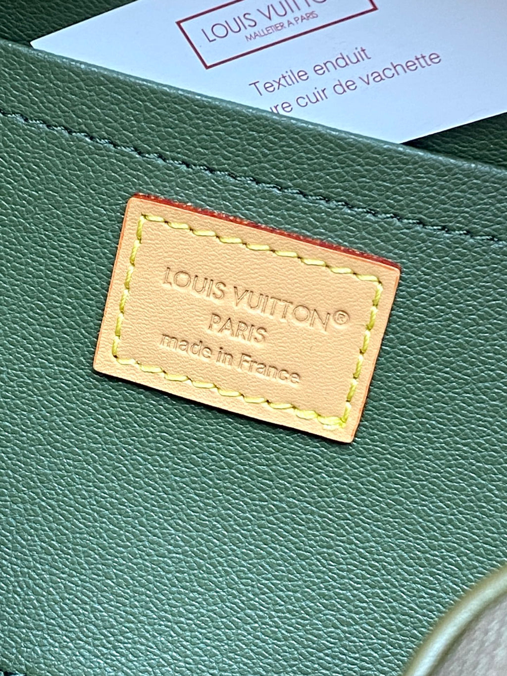 Green leather logo patch with yellow stitching on an olive bag panel