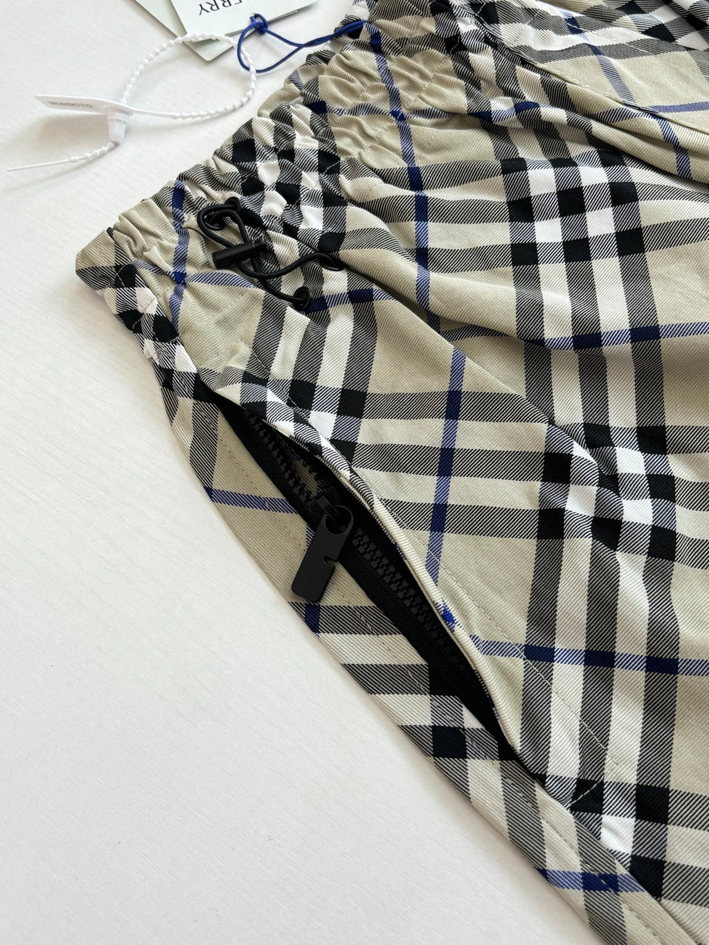 Plaid pants fabric close-up with black drawstring and zipper, beige with blue and black checks