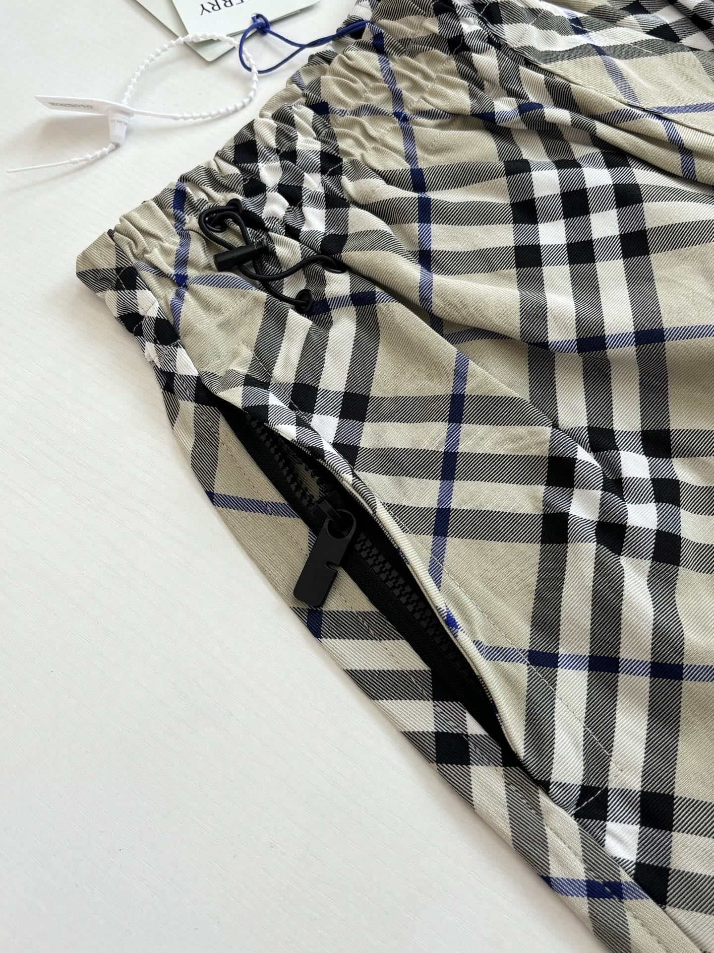 Plaid pants fabric close-up with black drawstring and zipper, beige with blue and black checks