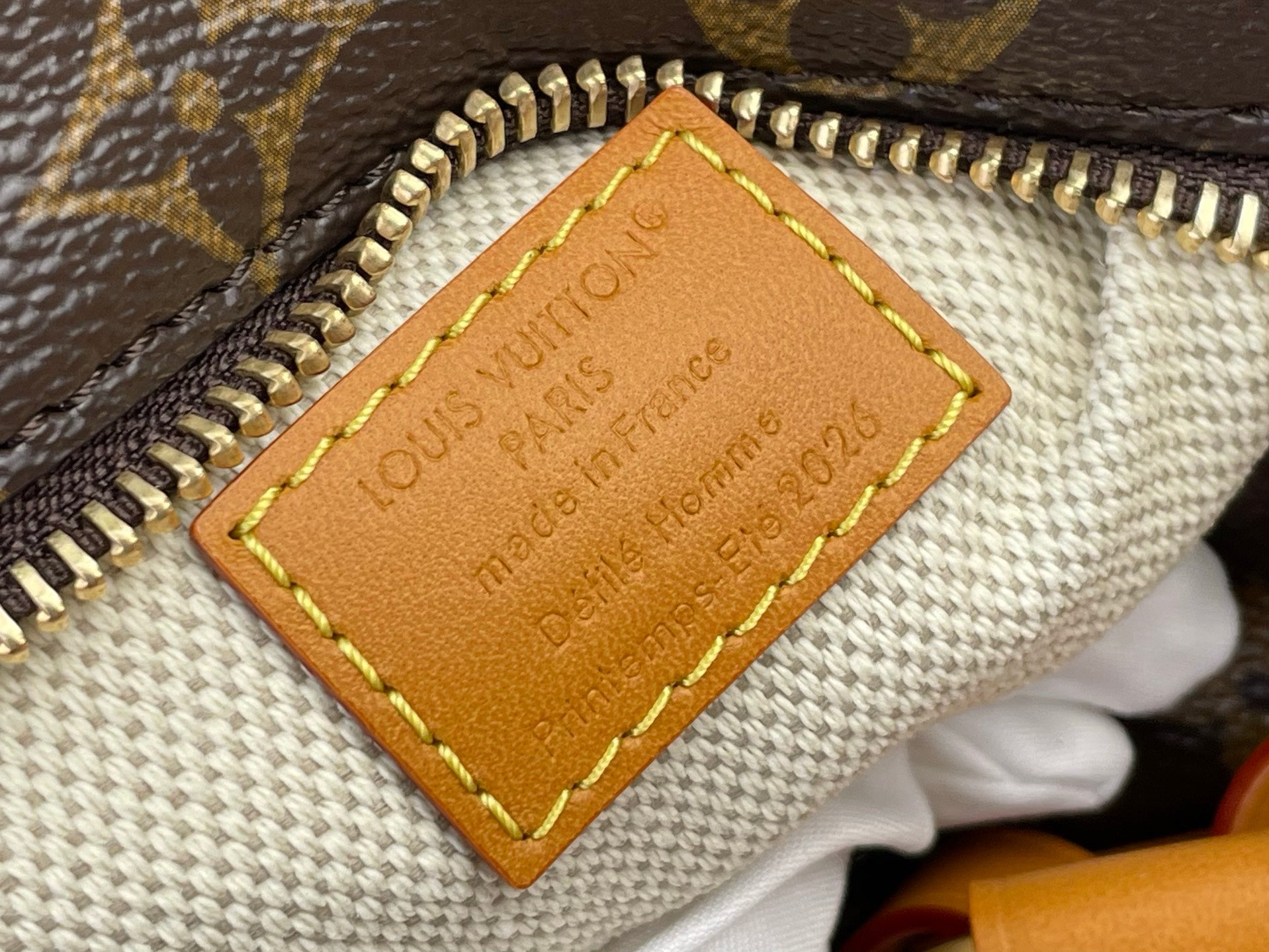 Tan leather inner label with embossed text and yellow edge stitching on monogram fabric