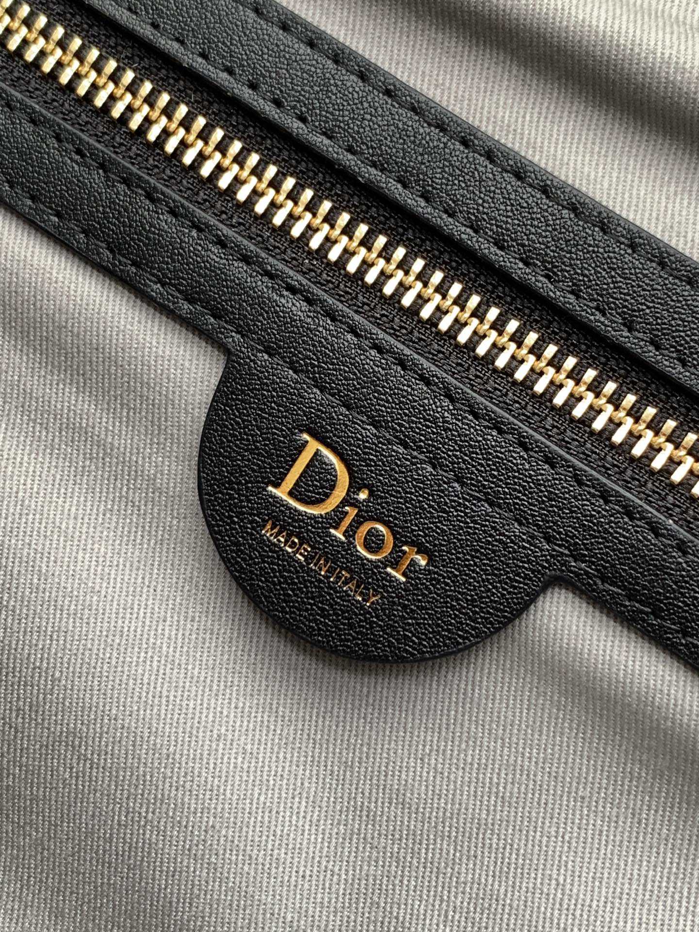 Dior bag zipper close-up with gold teeth on black leather, Dior logo on tab.