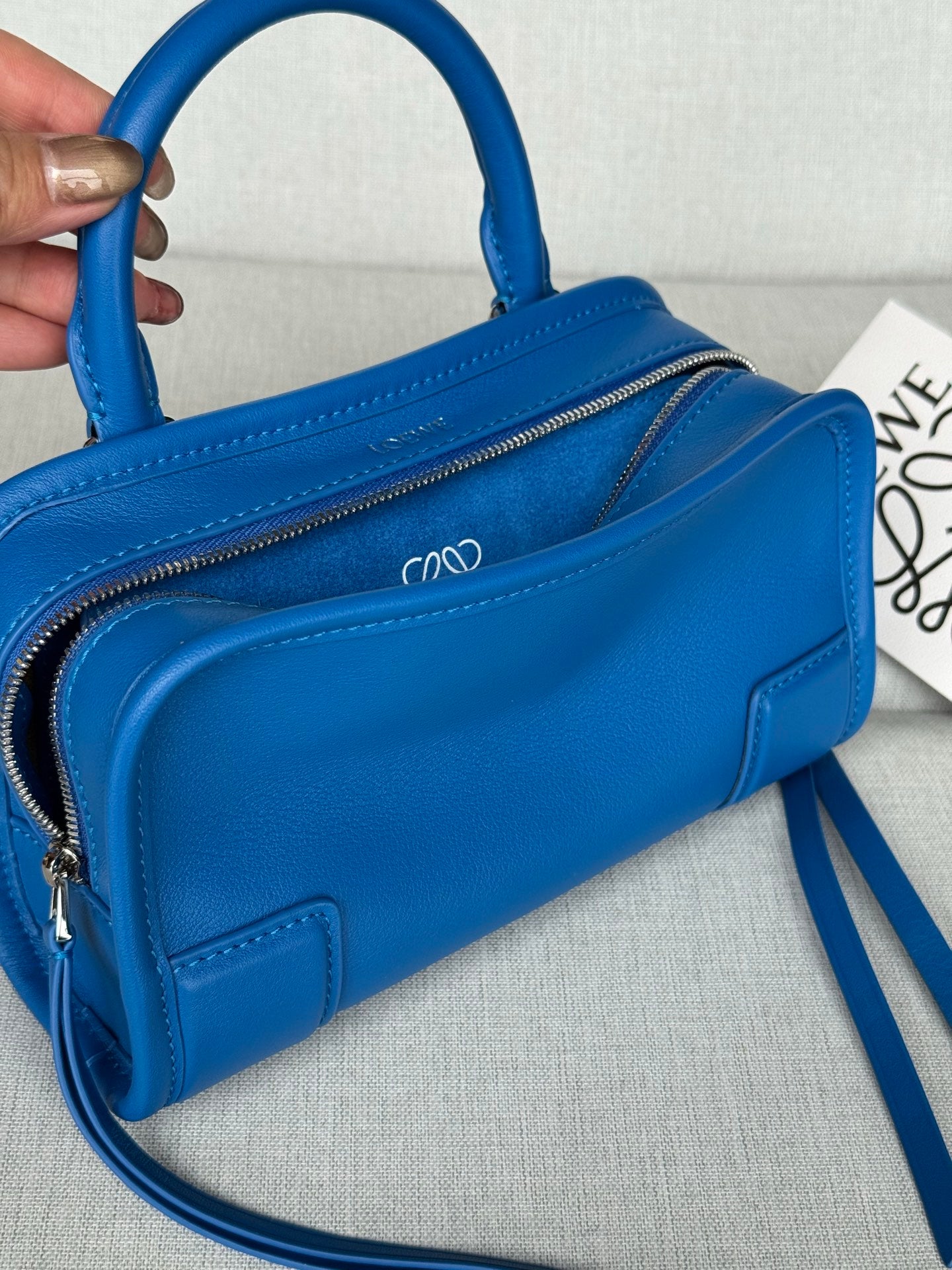 Blue leather bag with zip opening, front logo, and crossbody strap; hand holding handle