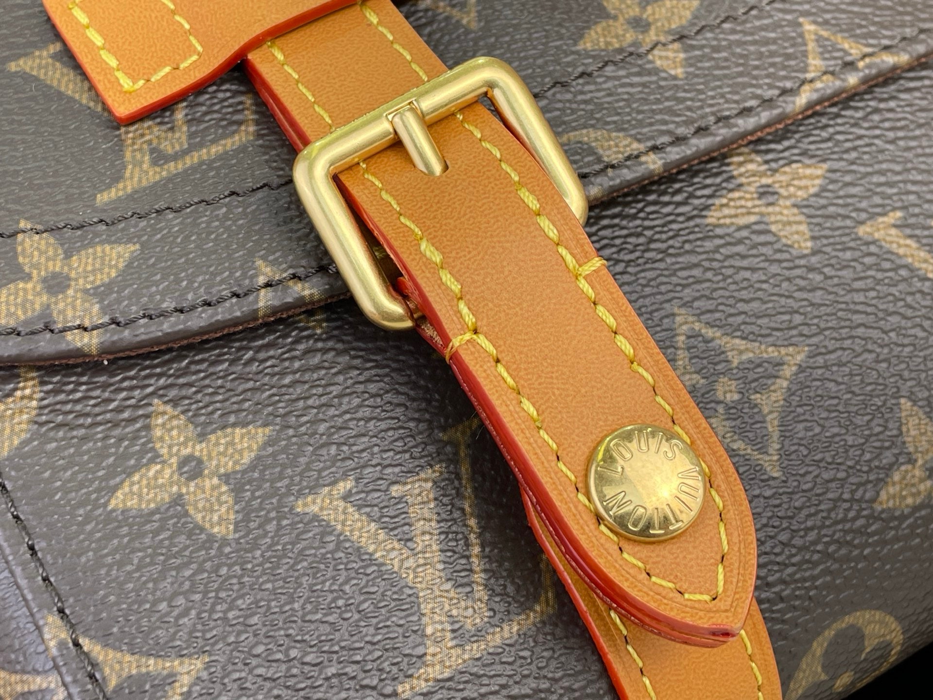 Monogram canvas strap close-up with tan leather, gold buckle, and red edge stitch