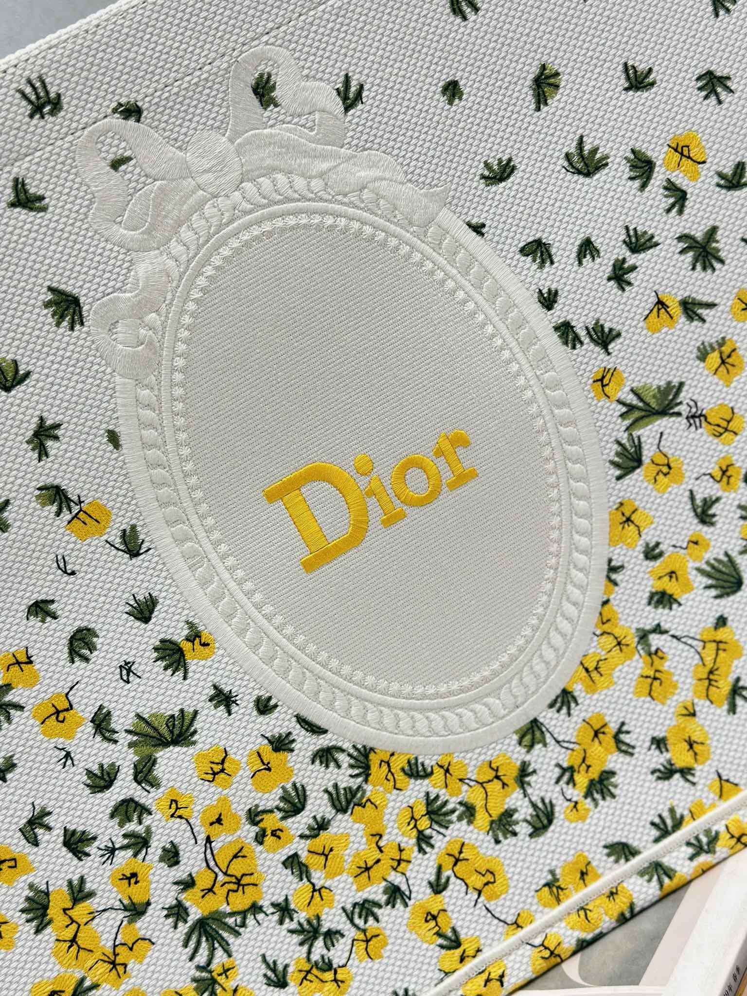 Dior oval logo patch on white tote bag fabric with yellow floral and cactus print.