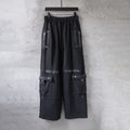 Cargo pants in dark fabric with elastic waistband and side utility pockets, zips, and straps