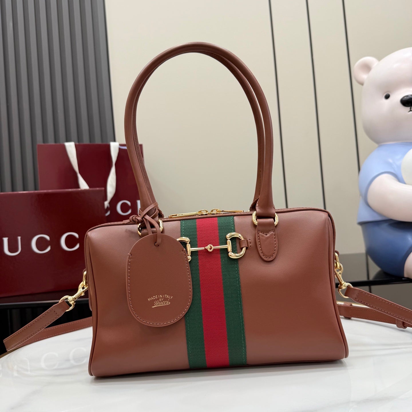 Brown leather handbag with green-and-red stripe, gold hardware, and hanging charm tag.