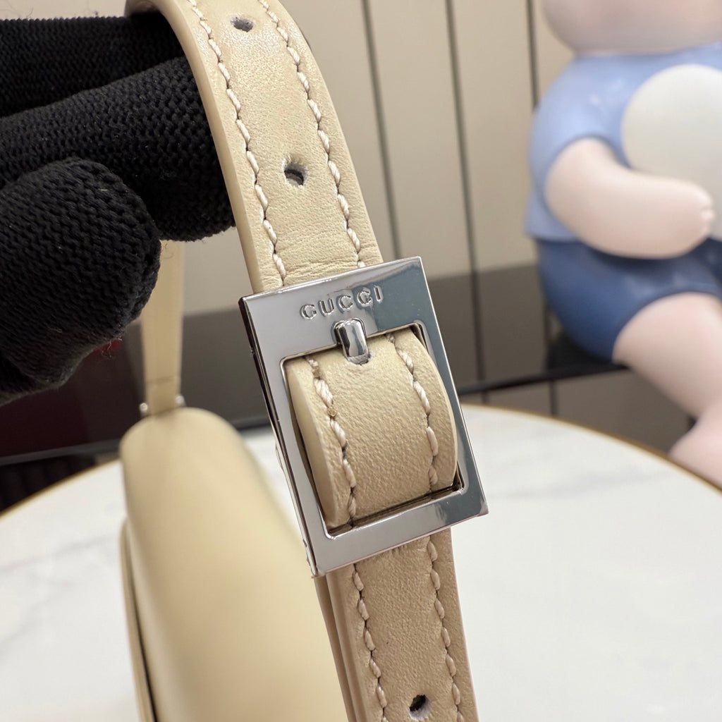 Gucci bag strap close-up in light beige with metal buckle and Gucci logo plate.