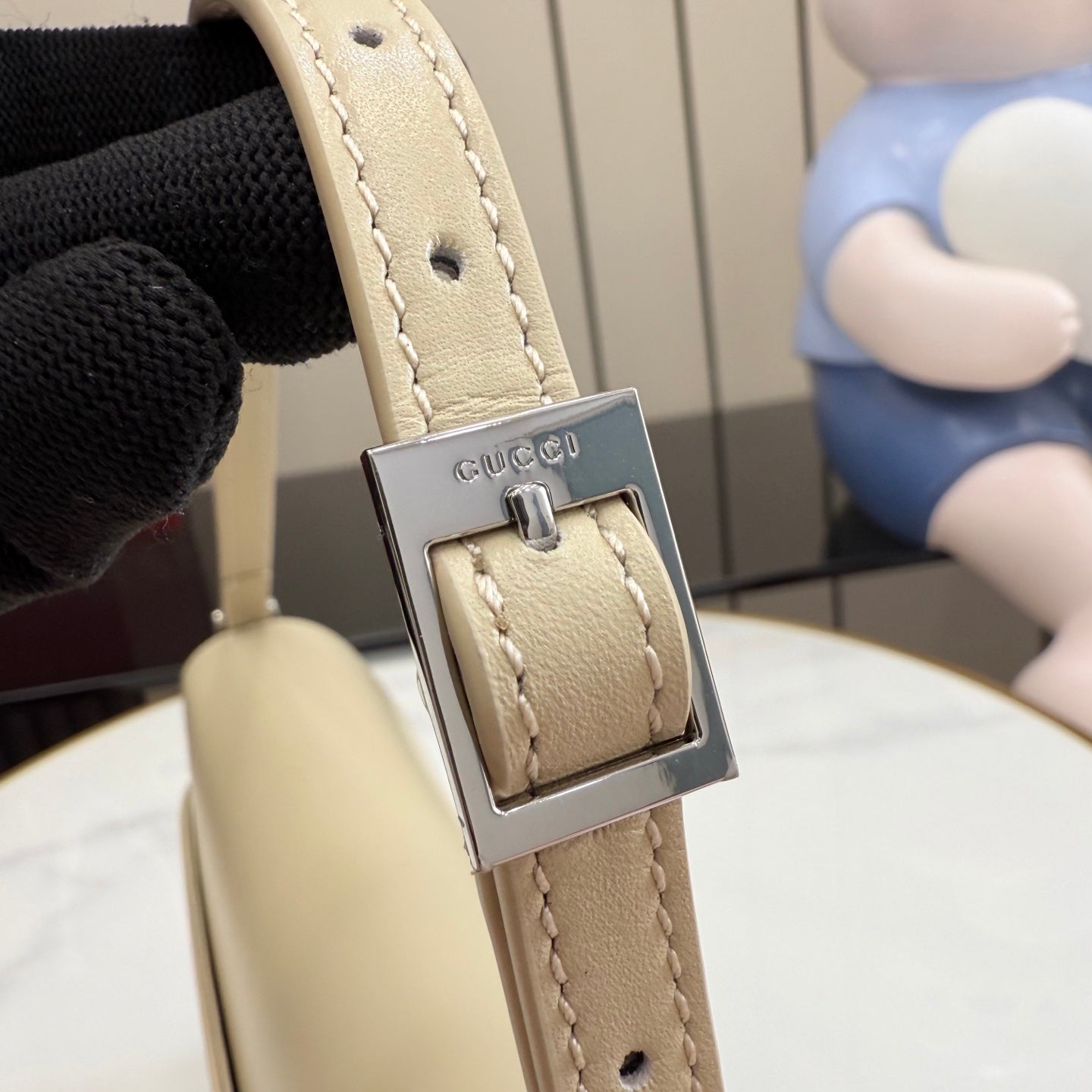 Gucci bag strap close-up in light beige with metal buckle and Gucci logo plate.