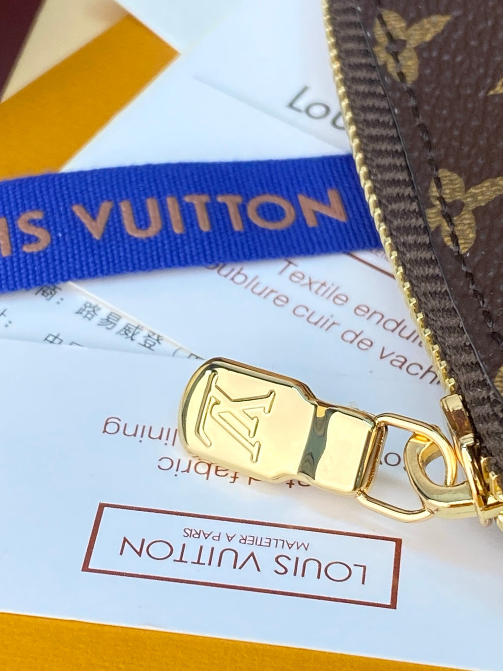 Gold zipper pull on a monogram pouch with blue strap and packaging nearby