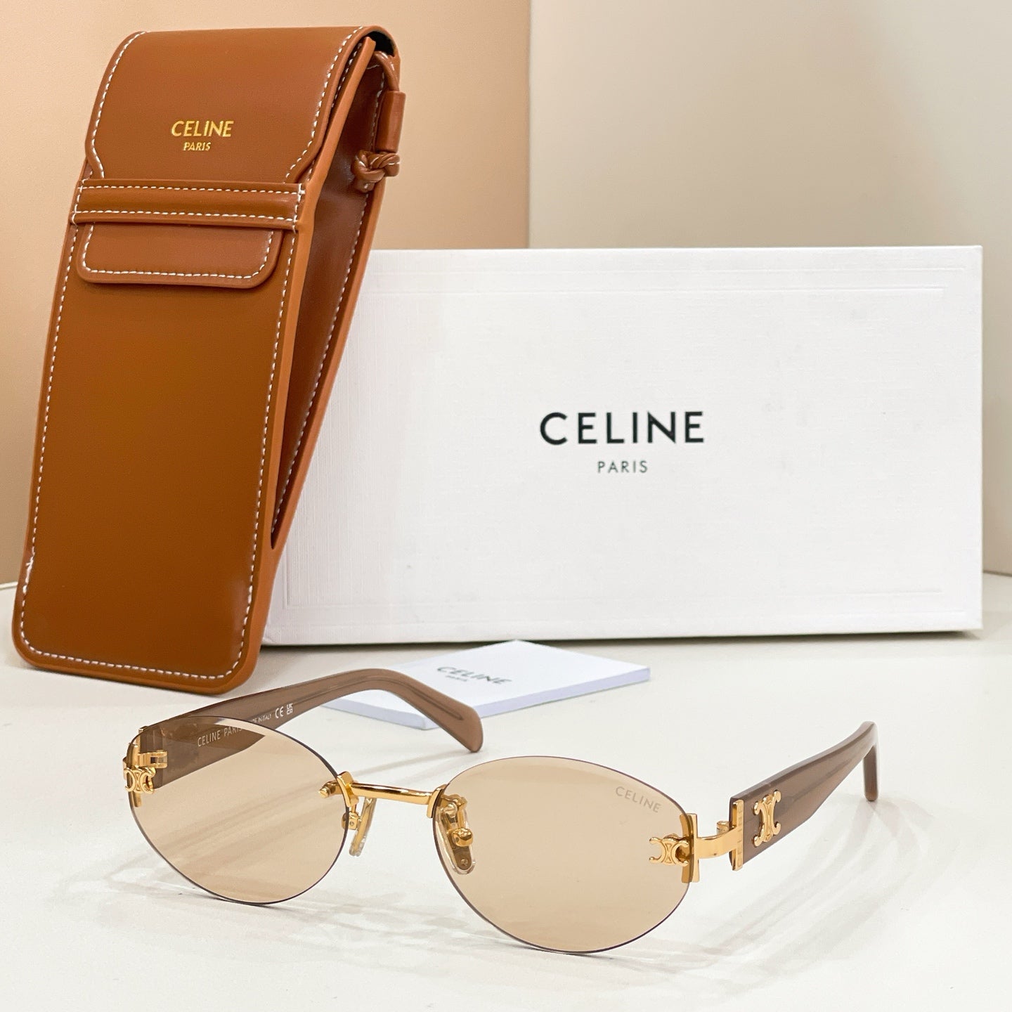 Cat-eye sunglasses with gold frame, light beige lenses, and brown temple arms with logo detail.