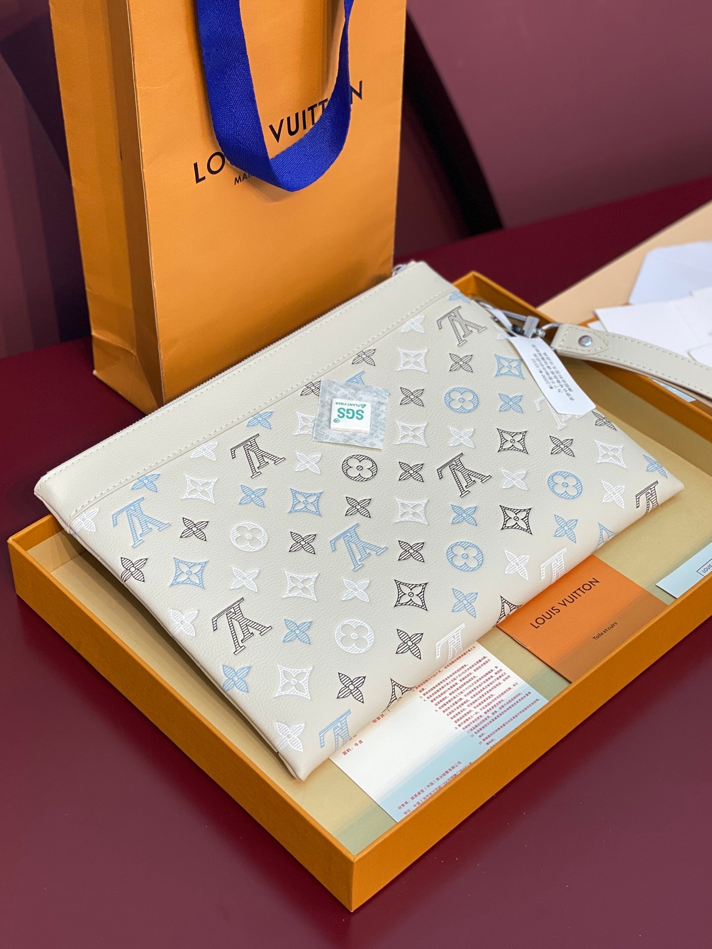 Small zip clutch bag in white with blue monogram pattern, shown on display box.