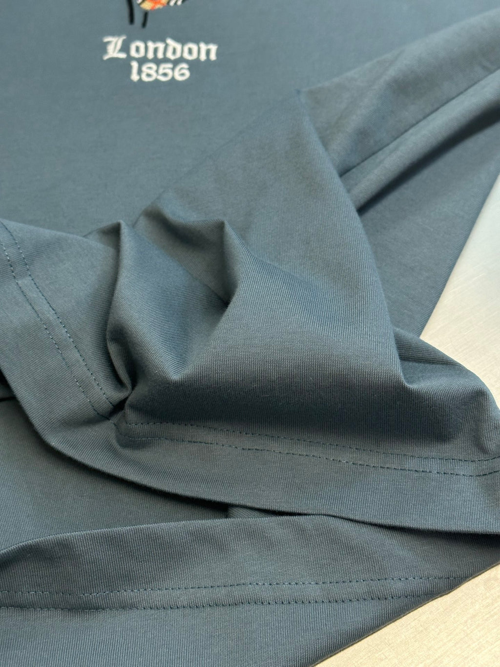 Blue T-shirt fabric close-up with stitched hem and partial “London 1856” text