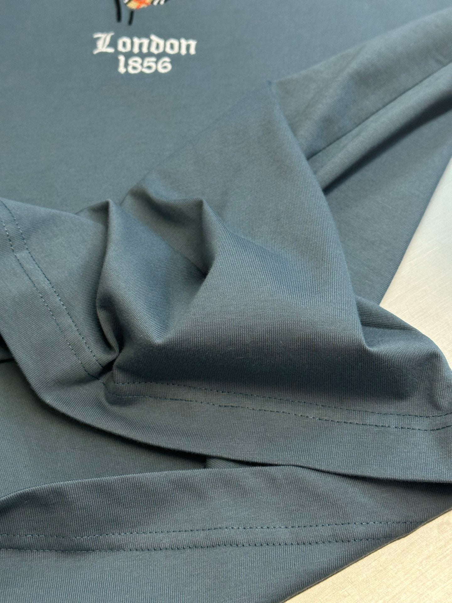 Blue T-shirt fabric close-up with stitched hem and partial “London 1856” text