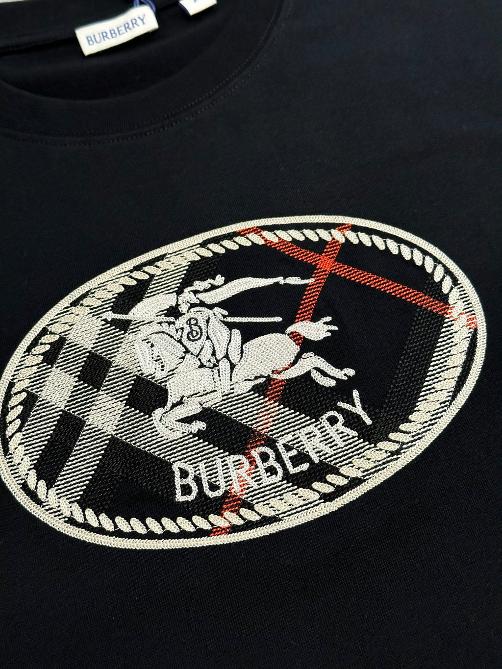 Black T-shirt chest graphic with oval Burberry logo and check pattern embroidery.