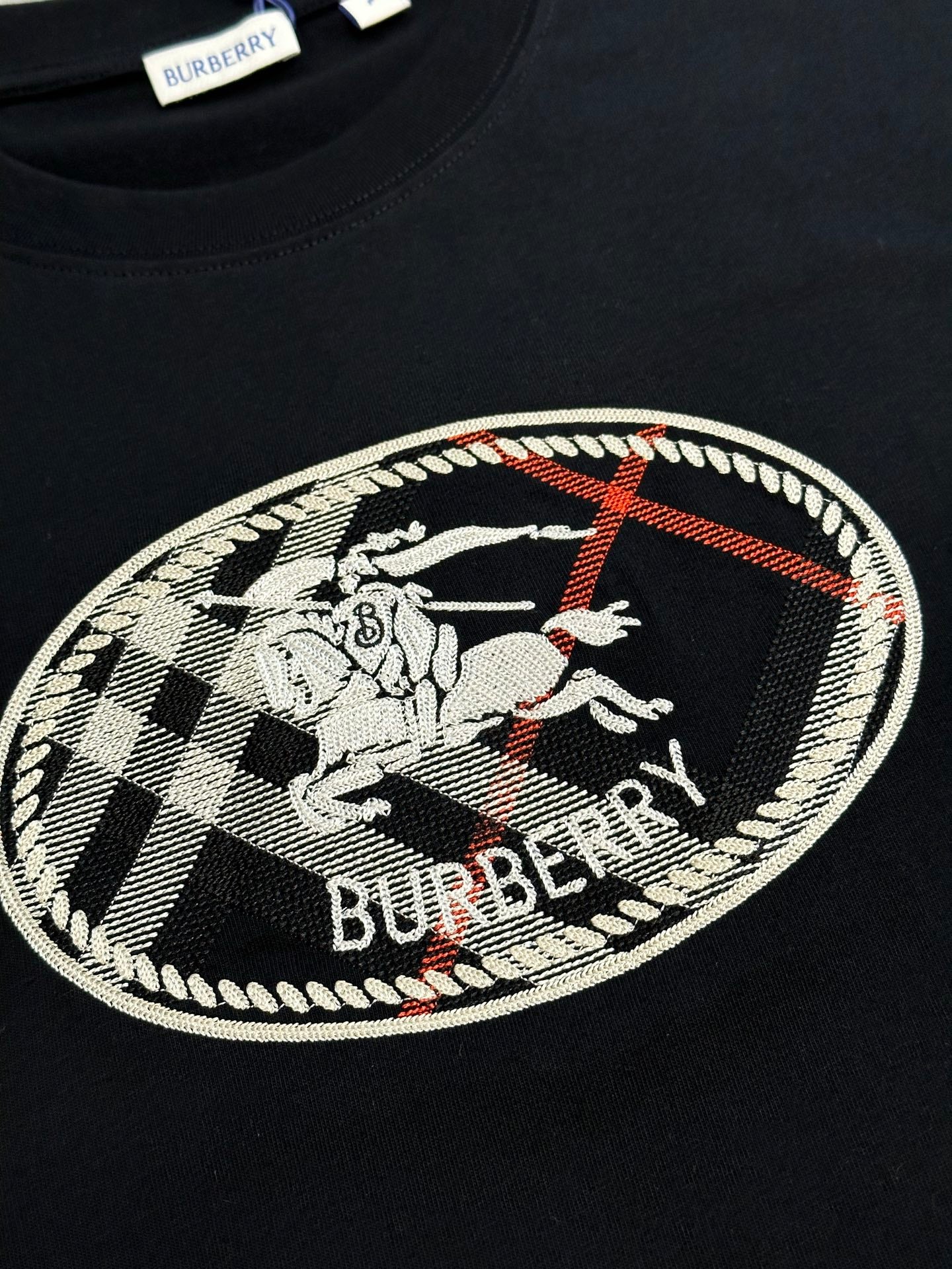 Black T-shirt chest graphic with oval Burberry logo and check pattern embroidery.
