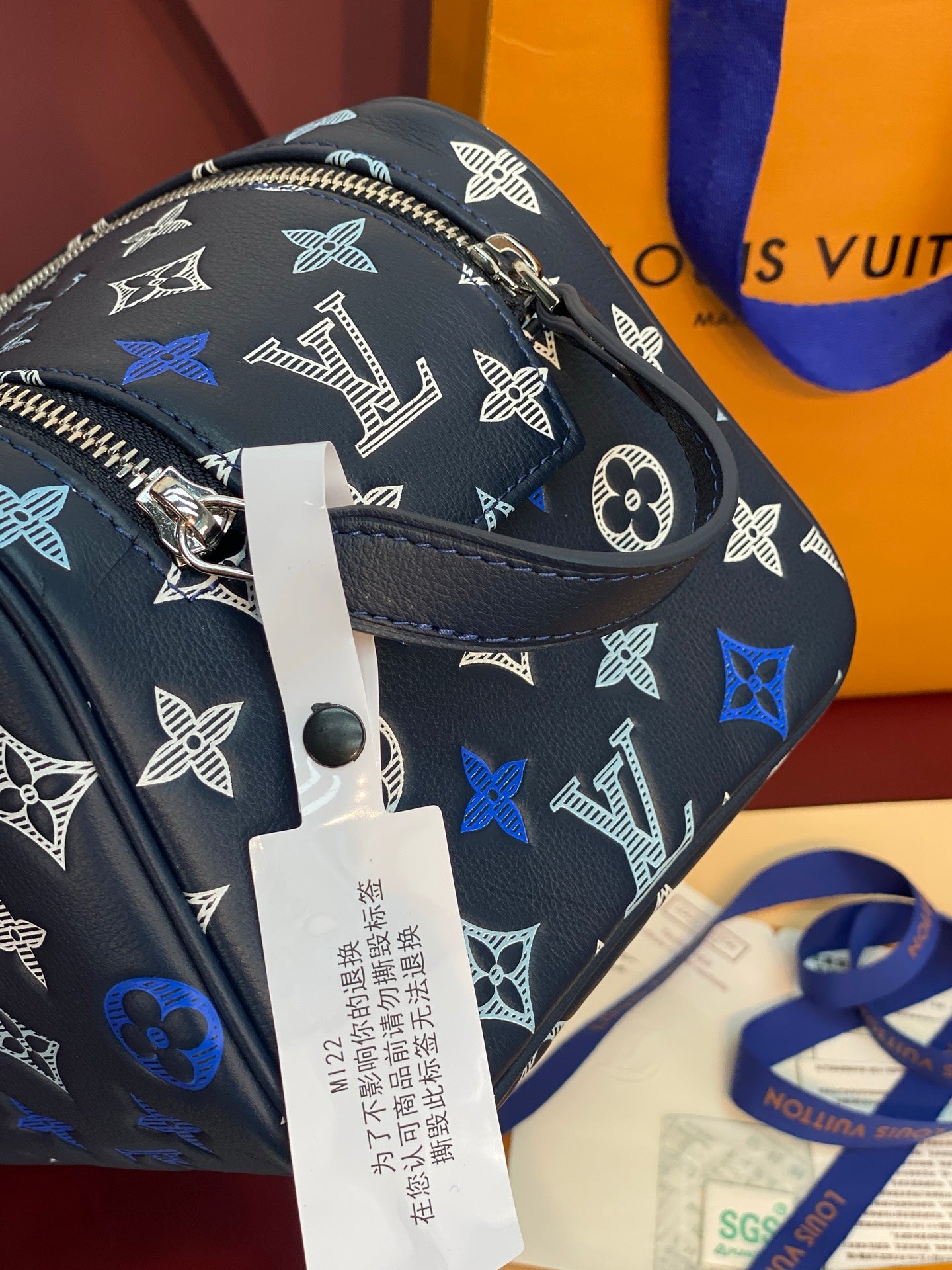 Louis Vuitton bag close-up: black-blue monogram, metal zipper pull, hanging authenticity tag.