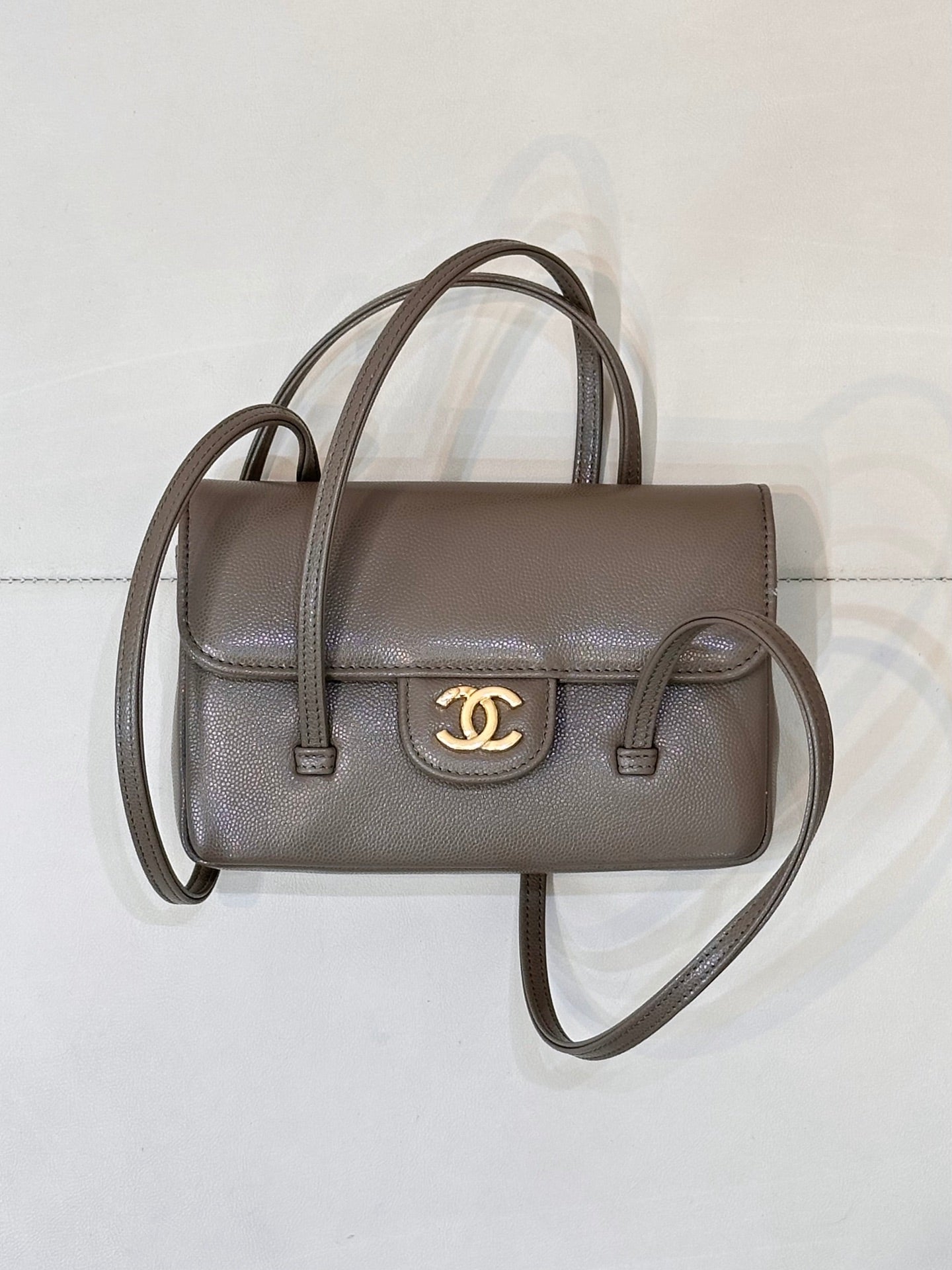 Chanel bag in taupe leather with two top handles, gold CC logo on front flap, and strap.