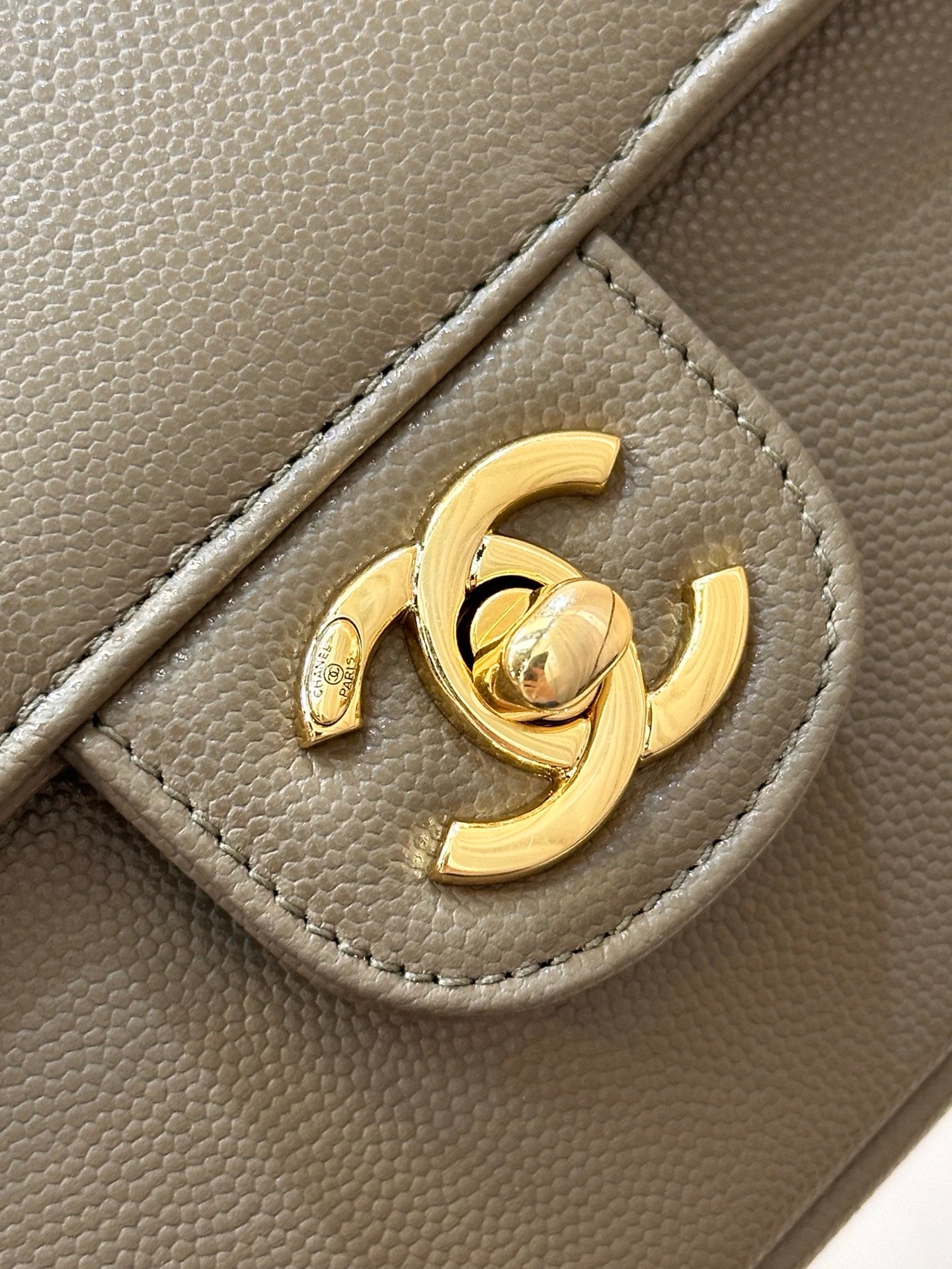 Gold CC logo detail on taupe Chanel bag hardware, with textured leather background.