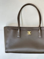 Taupe-brown leather tote bag close view, gold Chanel logo emblem on front, top handles