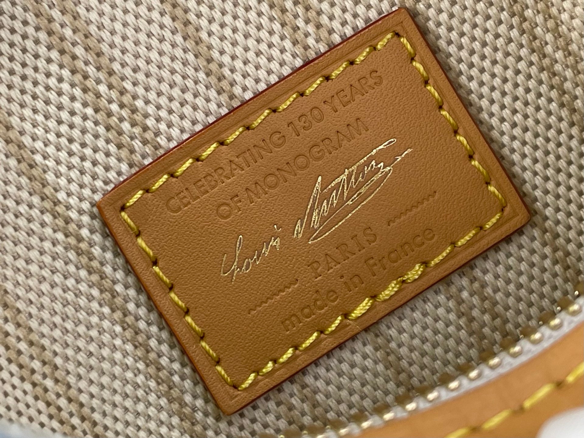 Tan leather brand patch with yellow stitching on light blue monogram canvas bag