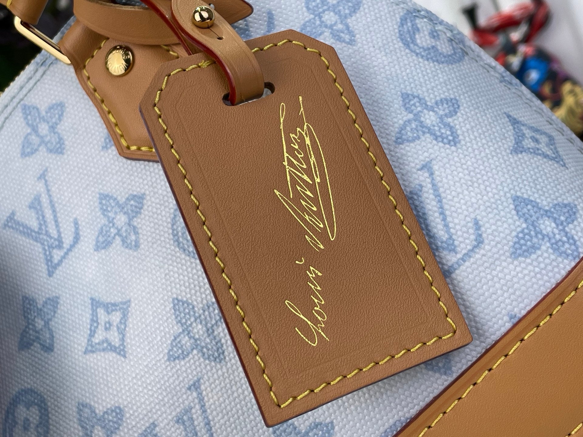 Tan leather luggage tag with stitched edges and gold signature on blue monogram fabric.