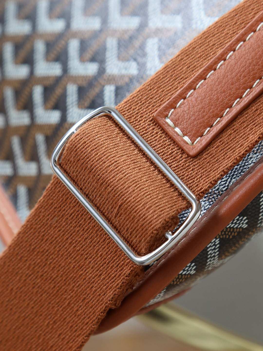 Goyard bag strap close-up with silver buckle and brown leather edging on monogram fabric.